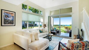 A spacious living room with a white sofa, a colorful chair, and a view of the outdoors through large sliding glass doors. - Ocean View - AC & Wifi - Walk to Beach & Shopping - Vrkcv (Kailua-Kona)
