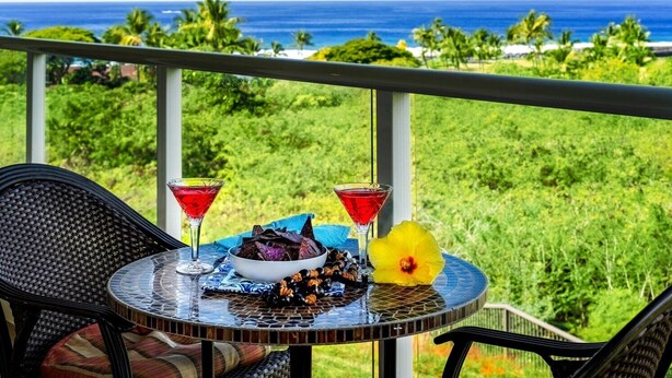 Een tafel met een schaal lekkernijen, twee martini glazen en een gele bloem op een balkon met uitzicht op een weelderig groon landschap en de oceaan.