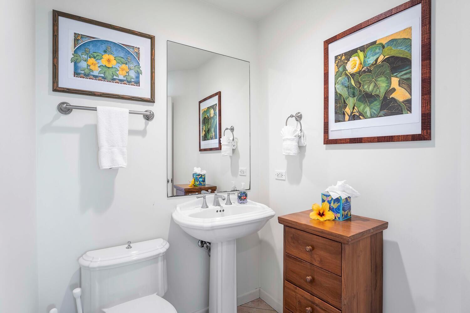 A bathroom with a toilet, sink, and vanity. There are framed pictures on the walls and a mirror above the sink.
