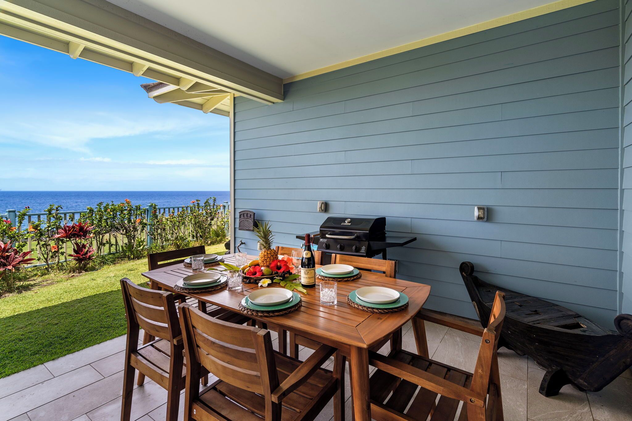 A wooden table set with plates, a bottle, and a fruit bowl, overlooking a garden with a view of the ocean.