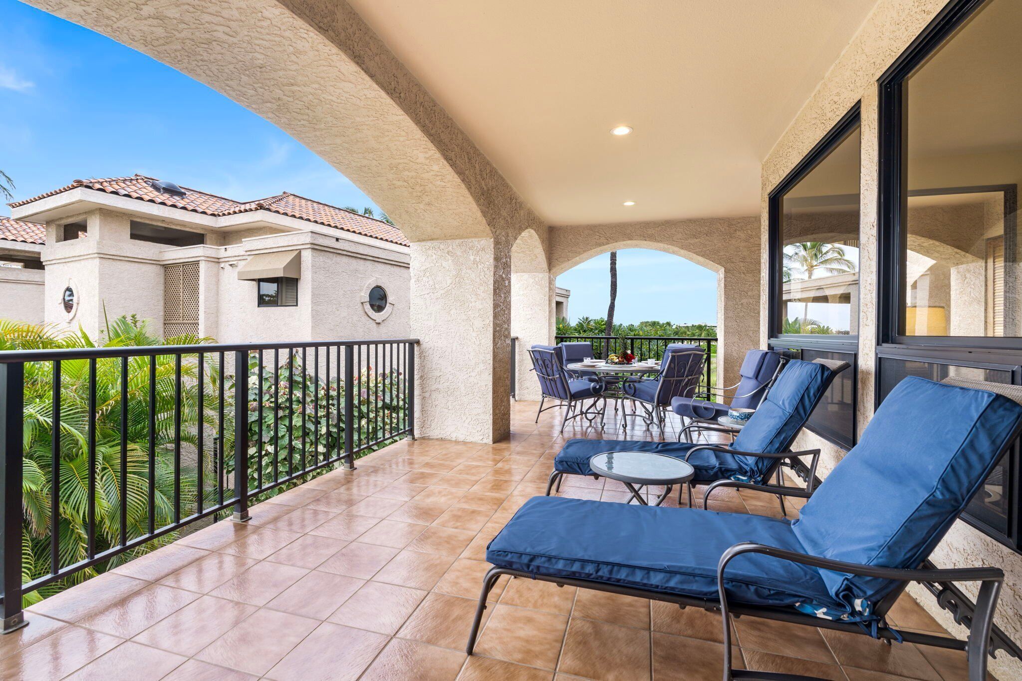 A covered outdoor area with lounge chairs and a table, surrounded by a railing and a view of buildings and greenery.