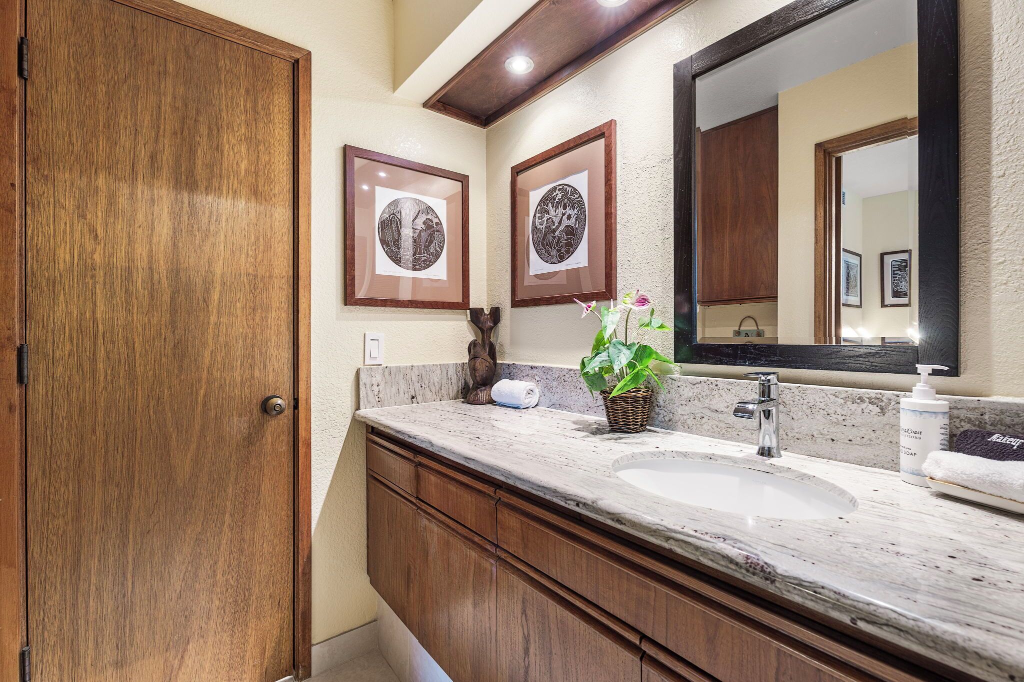 A bathroom with a wooden door, a marble sink, a large mirror, and two framed artworks on the wall.