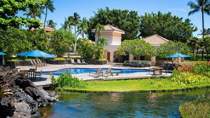 A pool area with lounge chairs and umbrellas, surrounded by tropical vegetation and a small water feature. - Relaxing Resort Living - Free Concierge - AC! - Vrkcv (Waikoloa)