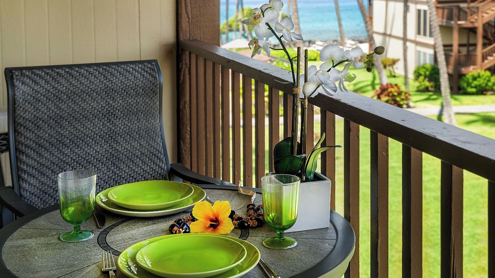 Un tavolo apparecchiato con piatti e bicchieri verdi, una pianta in vaso e un fiore giallo su un balcone con vista sull'oceano.
