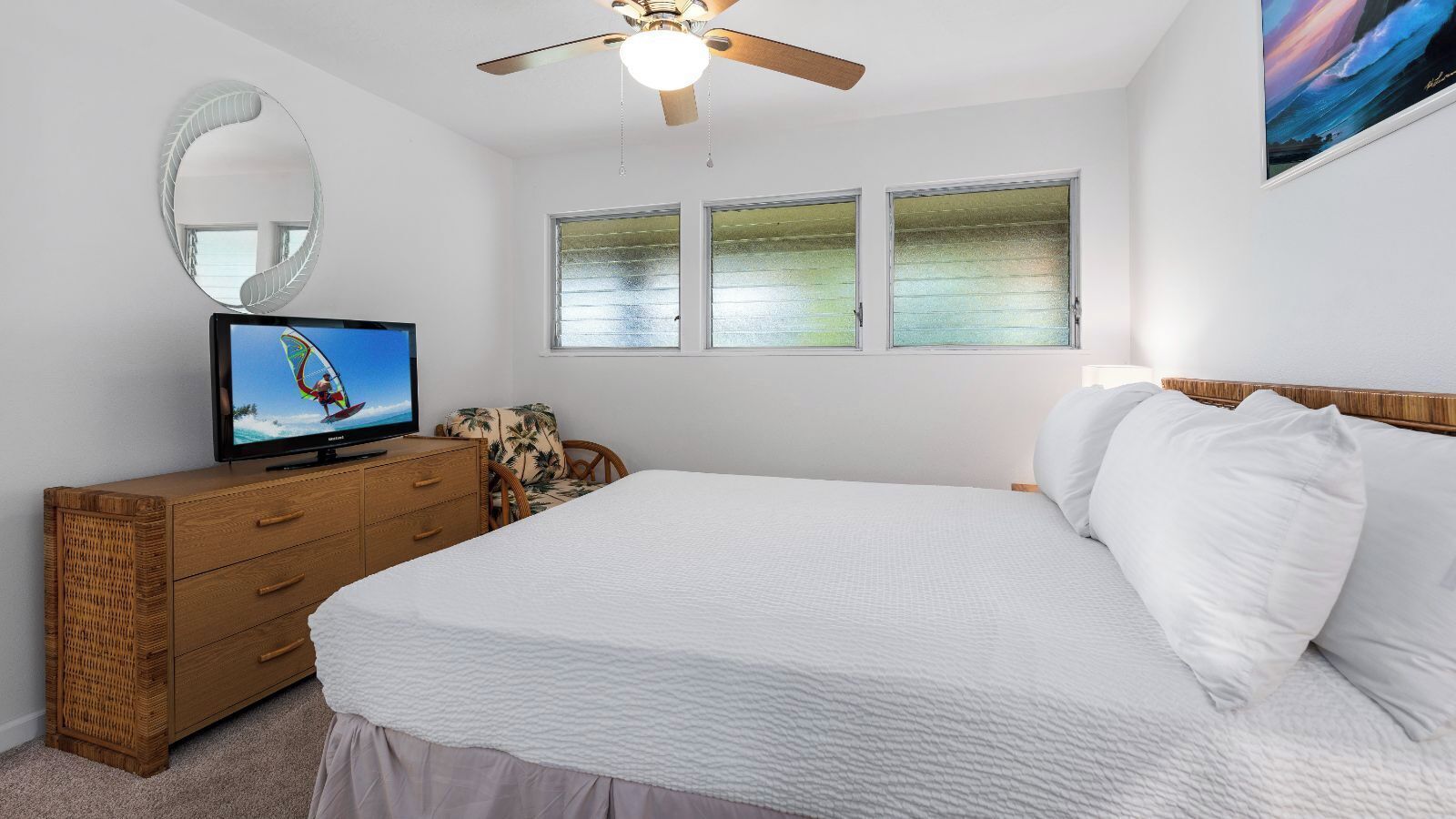 Ein Schlafzimmer mit einem Bett, einem Fernseher auf einer Holzkommode, einem Deckenventilator und drei Fenstern.