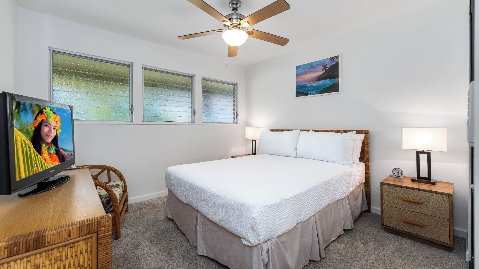 A bedroom with a bed, a television, a ceiling fan, a nightstand, and a window with a view of the ocean.