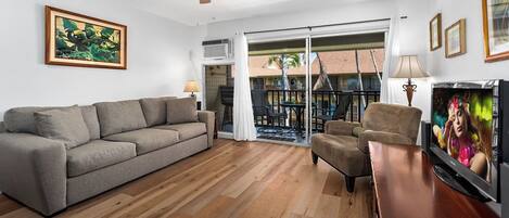 Ein Wohnzimmer mit grauem Sofa, einem Holztisch und einem Fernseher. Eine Glastür öffnet sich zu einem Außenbereich mit Pool und Palmen.
