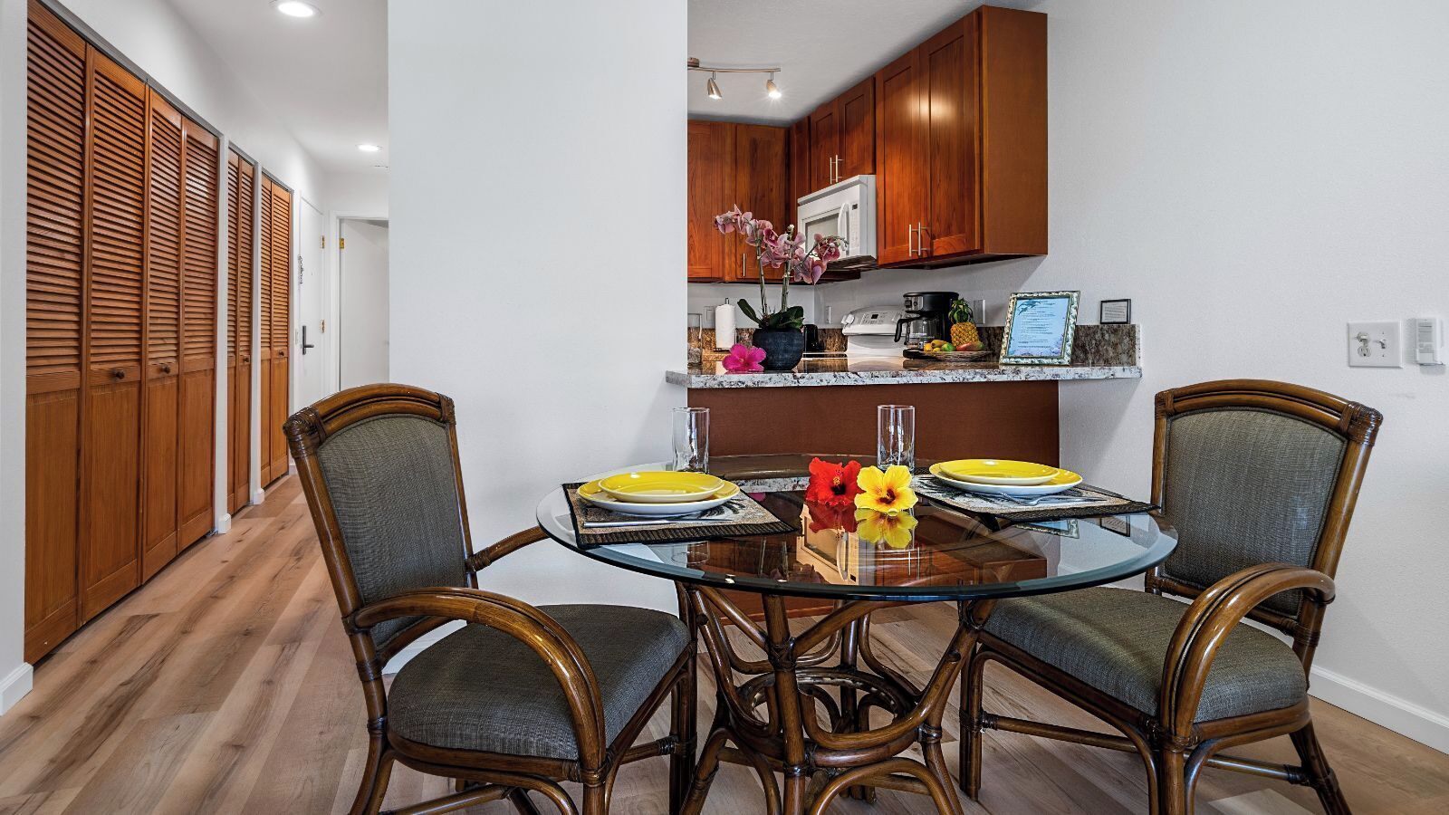 A dining area with a round glass table, two chairs, and a kitchen in the background.