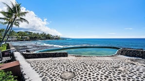 Un área empaveda de piedra con una piscina, palmeras y vista al océano.