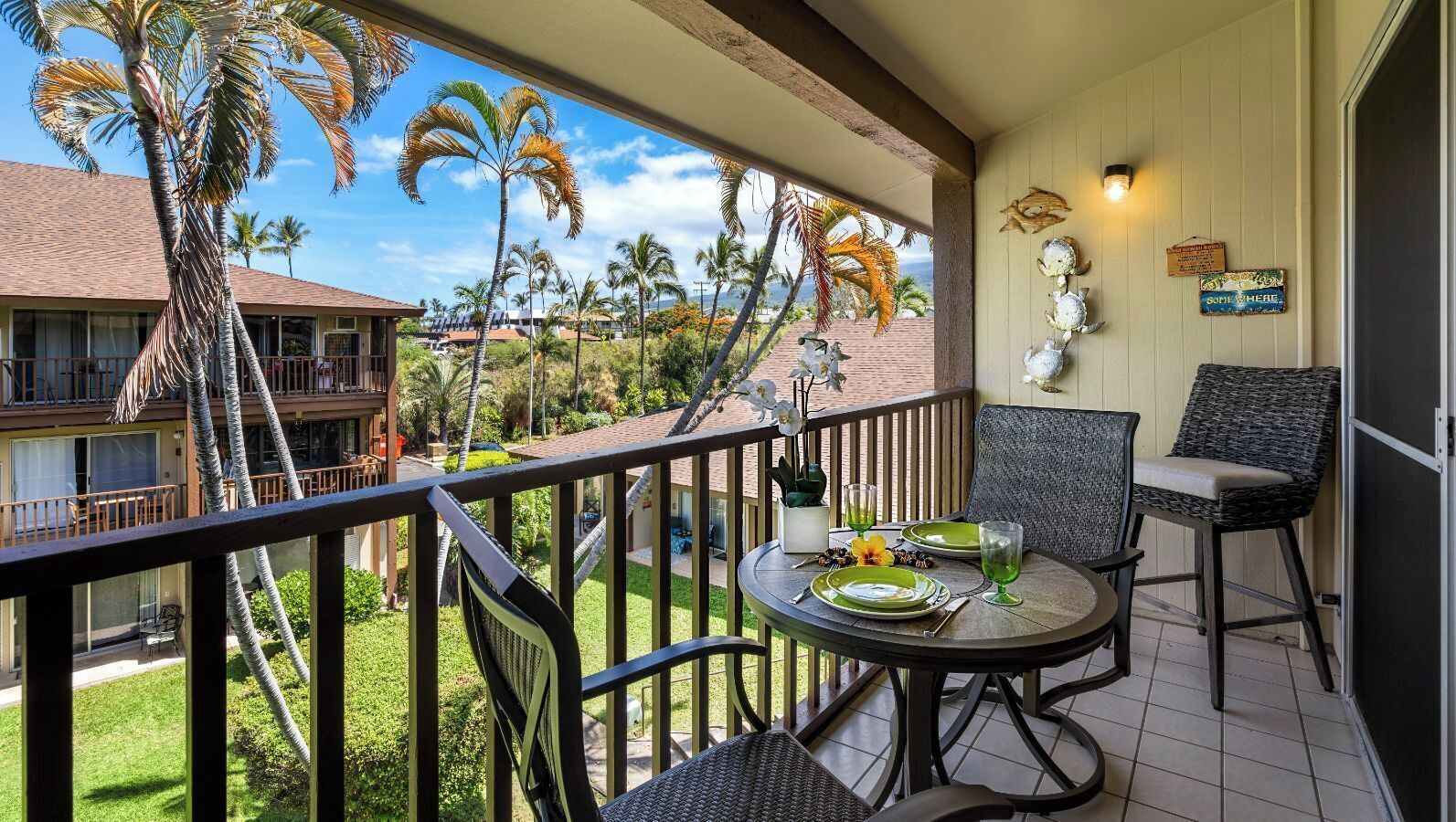 A balcony with a table set for two, overlooking a tropical landscape with palm trees and a view of a building across the street.