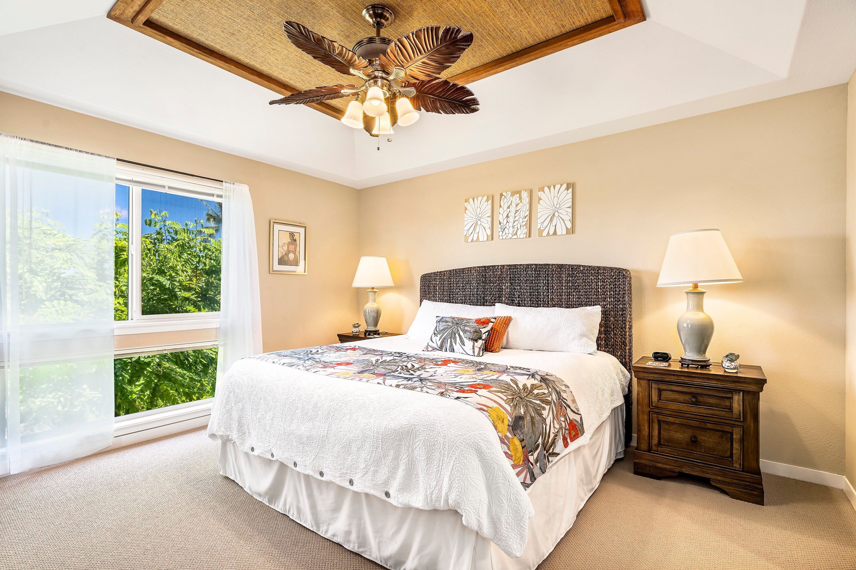 A bedroom with a large bed, two bedside tables with lamps, a ceiling fan, and a view of greenery through a large window.