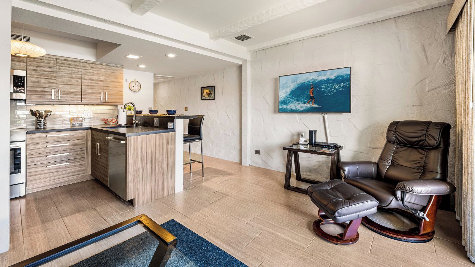 Een moderne keuken met houten kastjes en een centraal eiland, een woonruimte met een relaxfauteuil en een televisie, en een marmeren wand met een schilderij.