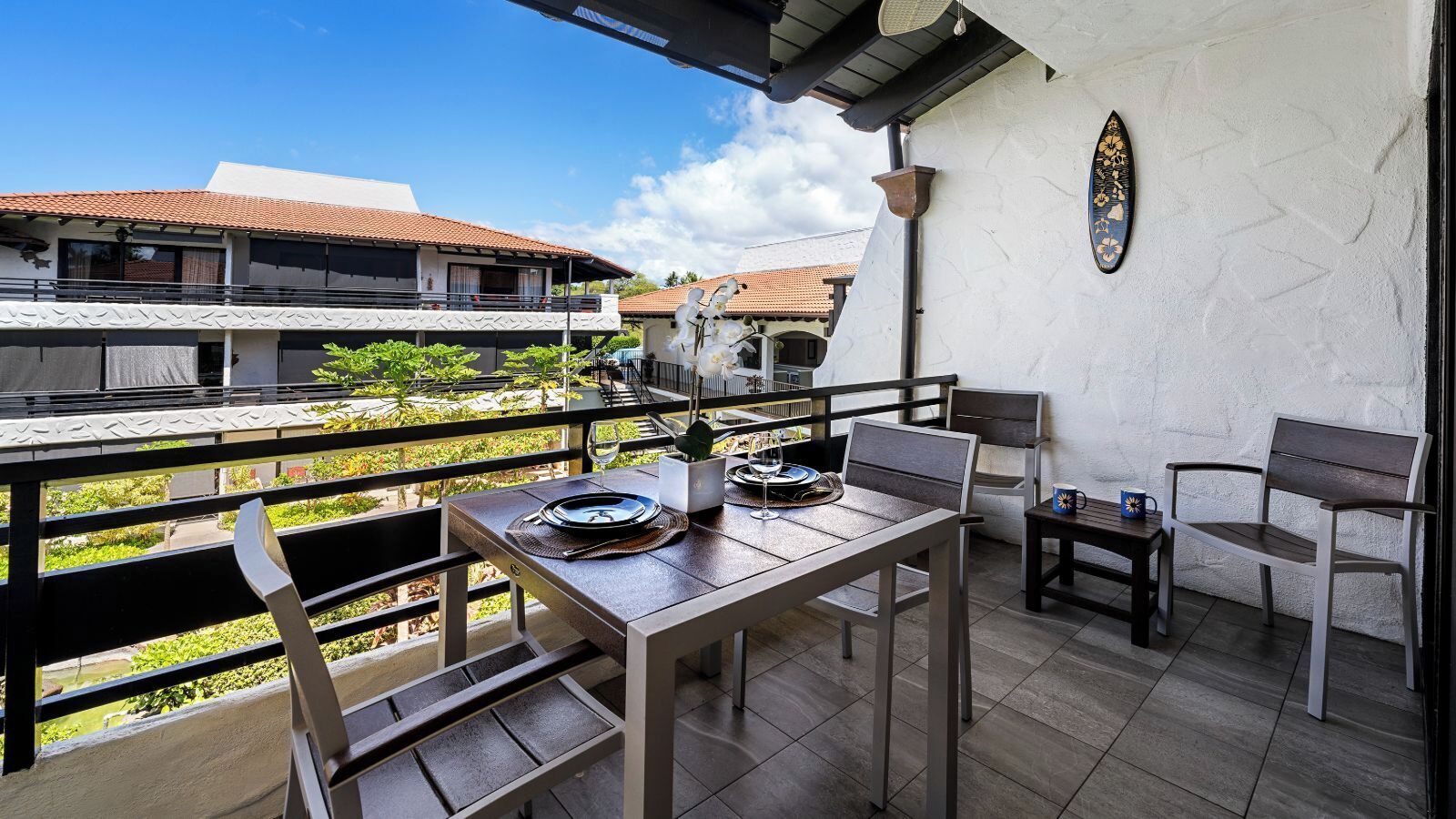 A balcony with a table set for two, chairs, a small table with a mug, and a decorative surfboard on the wall.