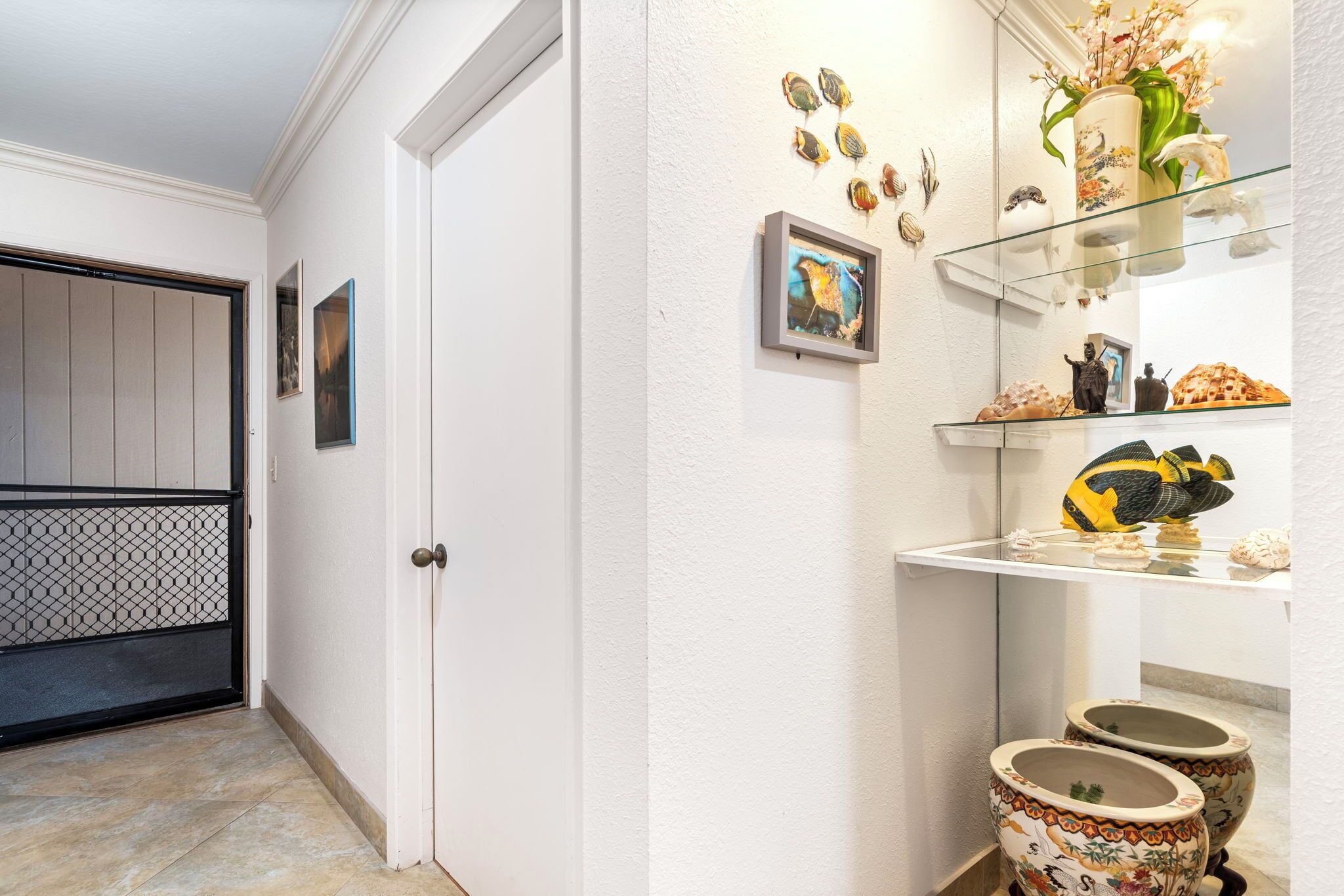 Un couloir avec une porte blanche, une étagère en verre où sont exposés des objets décoratifs et un sol carrelé.