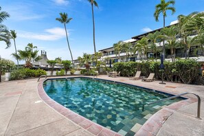 A swimming pool with a tiled edge, surrounded by palm trees and lounge chairs, with a building in the background.