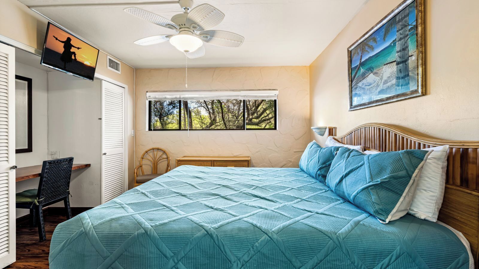 Ein Schlafzimmer mit Bett, Deckenventilator, Fernseher, Fenster mit Blick auf Bäume und einem Bild an der Wand.