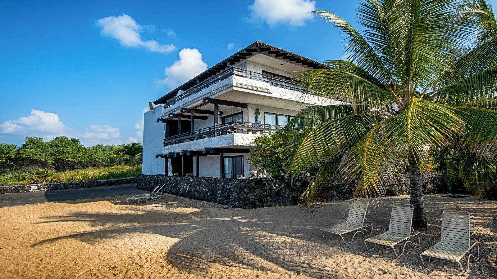 A two-story beachfront property with a balcony, a stone wall, and a palm tree.