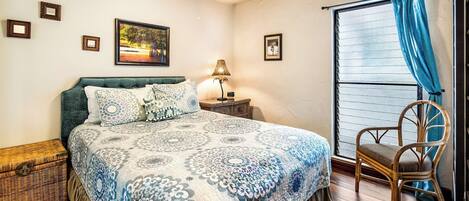 A bedroom with a bed, a nightstand, a lamp, a wicker chest, a chair, and a window with curtains.