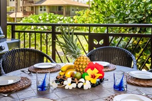 A table set outdoors with a tropical fruit arrangement, including pineapple, bananas, and mangoes, and a hibiscus flower centerpiece.