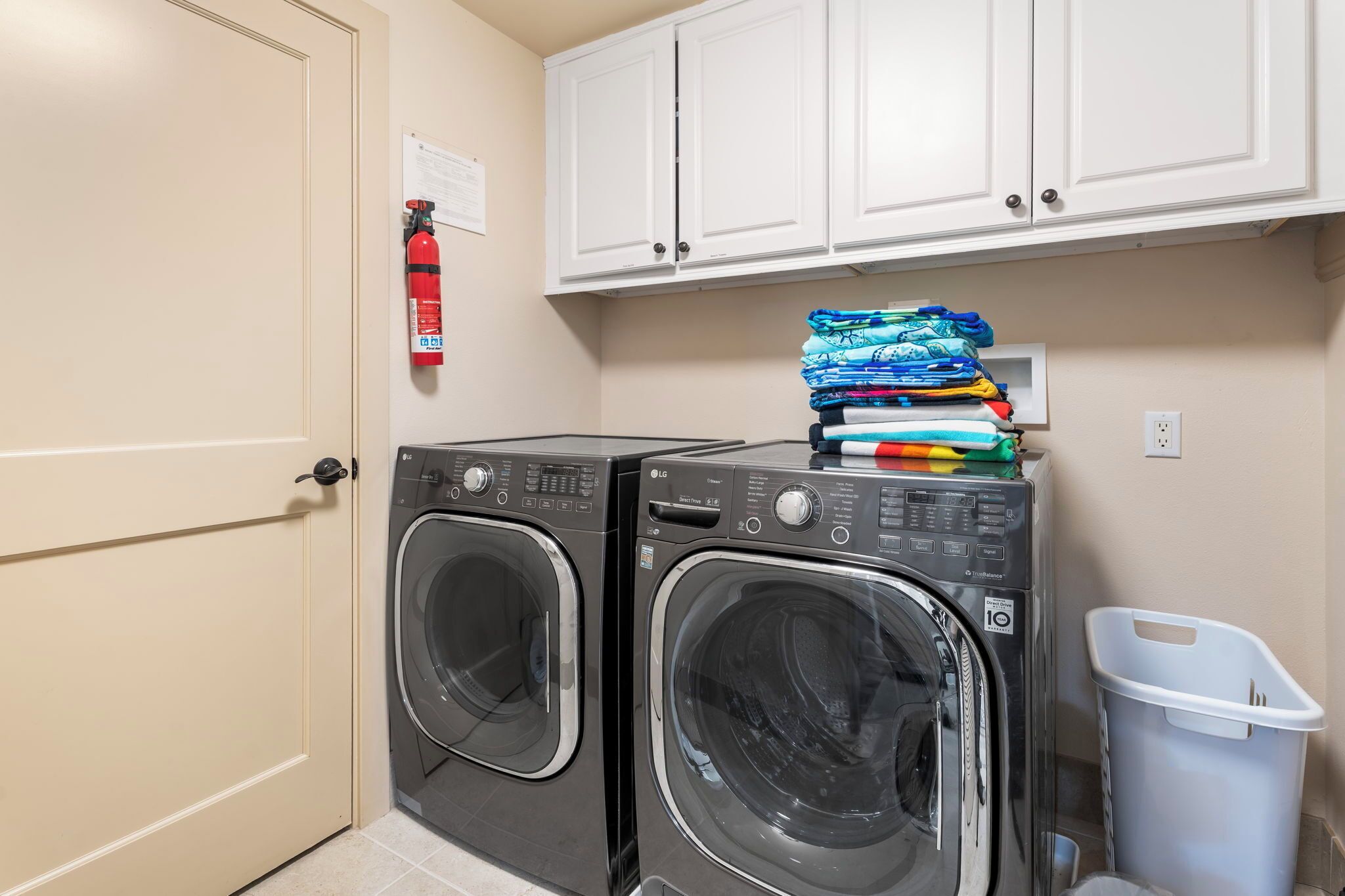 A laundry room with two stacked washing machines, a fire extinguisher, and a white trash bin.