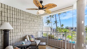 A balcony with a stone wall, a ceiling fan, a table with chairs, and a view of palm trees and a pool. - Oceanview Kona Condo - Pool, Lanai, Walk to Town - Vrkcv (Kailua-Kona)