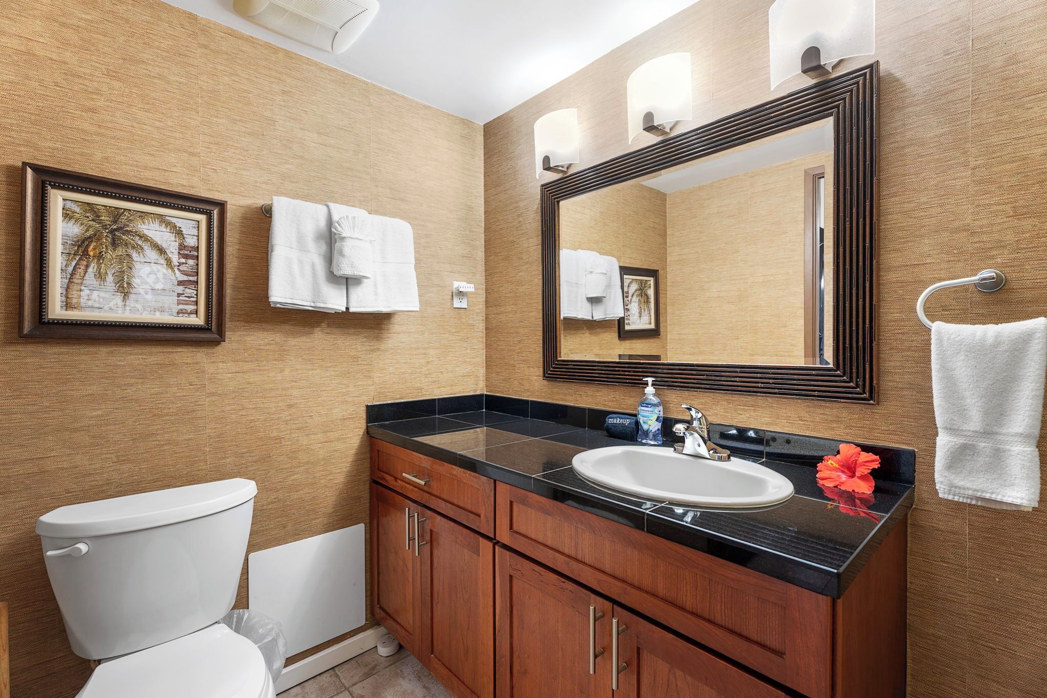 A bathroom with a toilet, sink, and mirror. There are towels hanging on the wall and a framed picture above the sink.