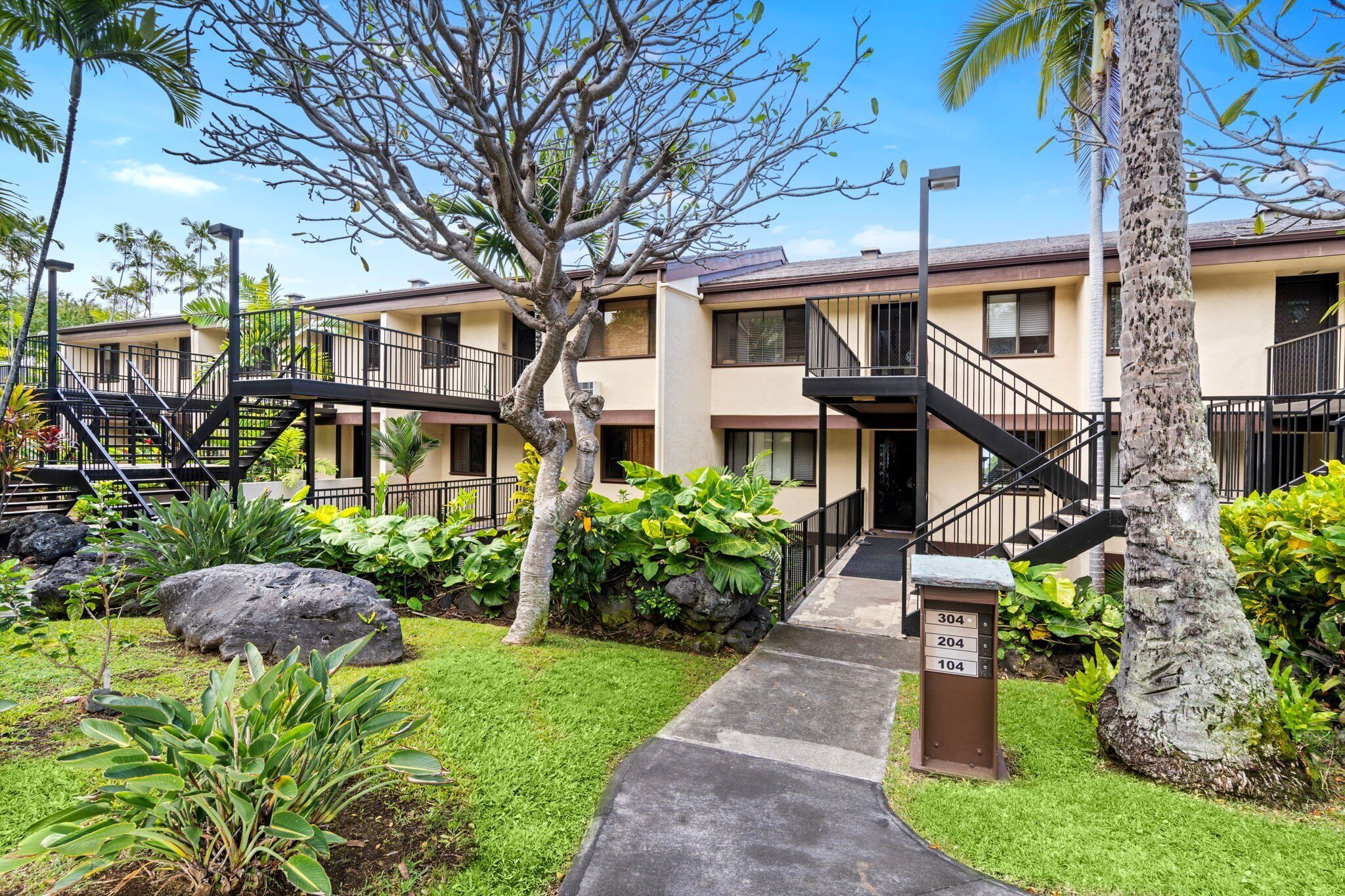 A two-story building with a central staircase, surrounded by lush greenery and palm trees.