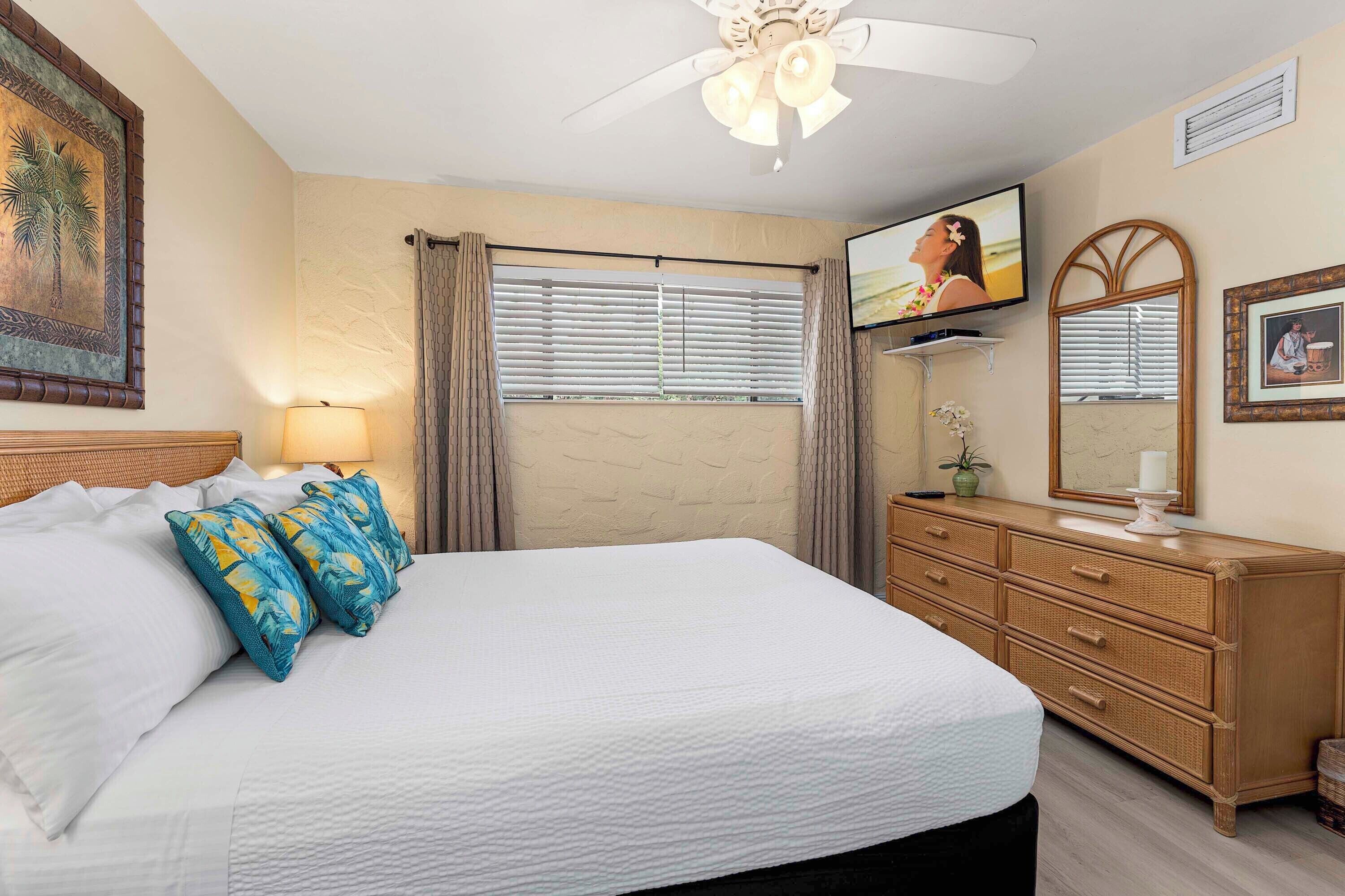A neatly made bed with white linens and blue pillows, a wooden dresser with drawers, a ceiling fan, a television on the wall, and a window with blinds.