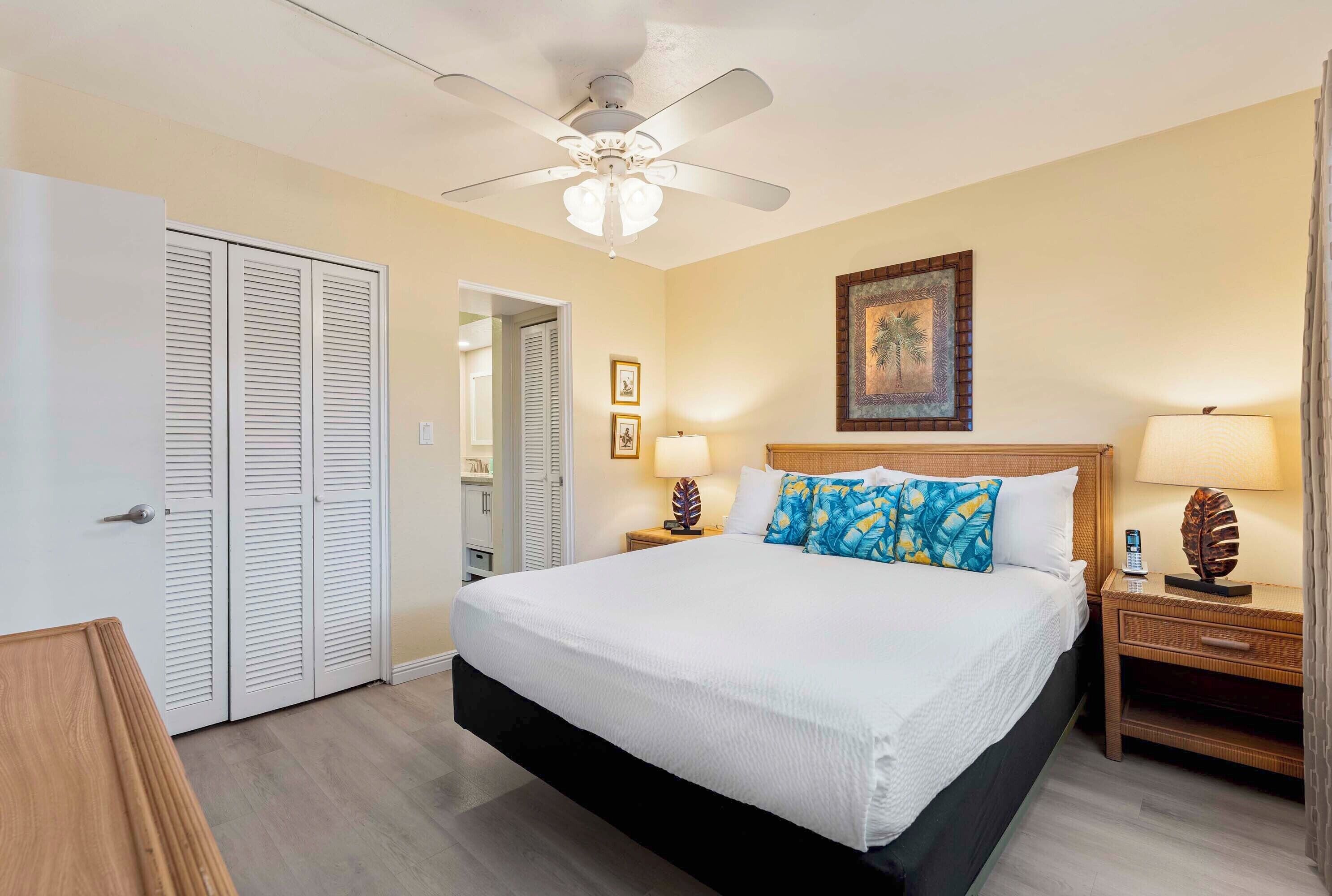 A hotel room with a large bed, a ceiling fan, a wooden nightstand, and a door with louvered shutters.