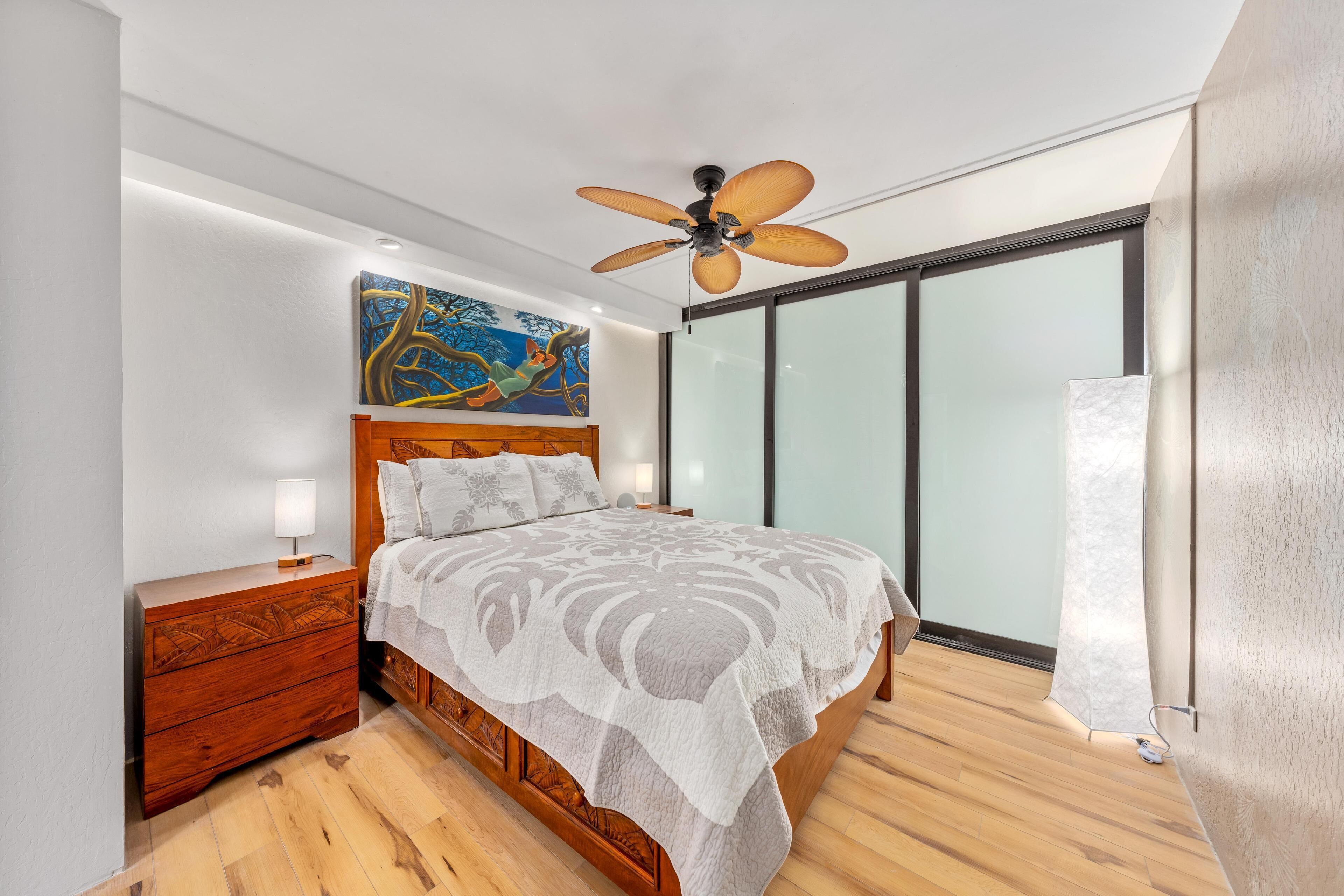 Een slaapkamer met een houten bed, een plafondventilator, een nachtkastje, een schilderij en een groot raam.