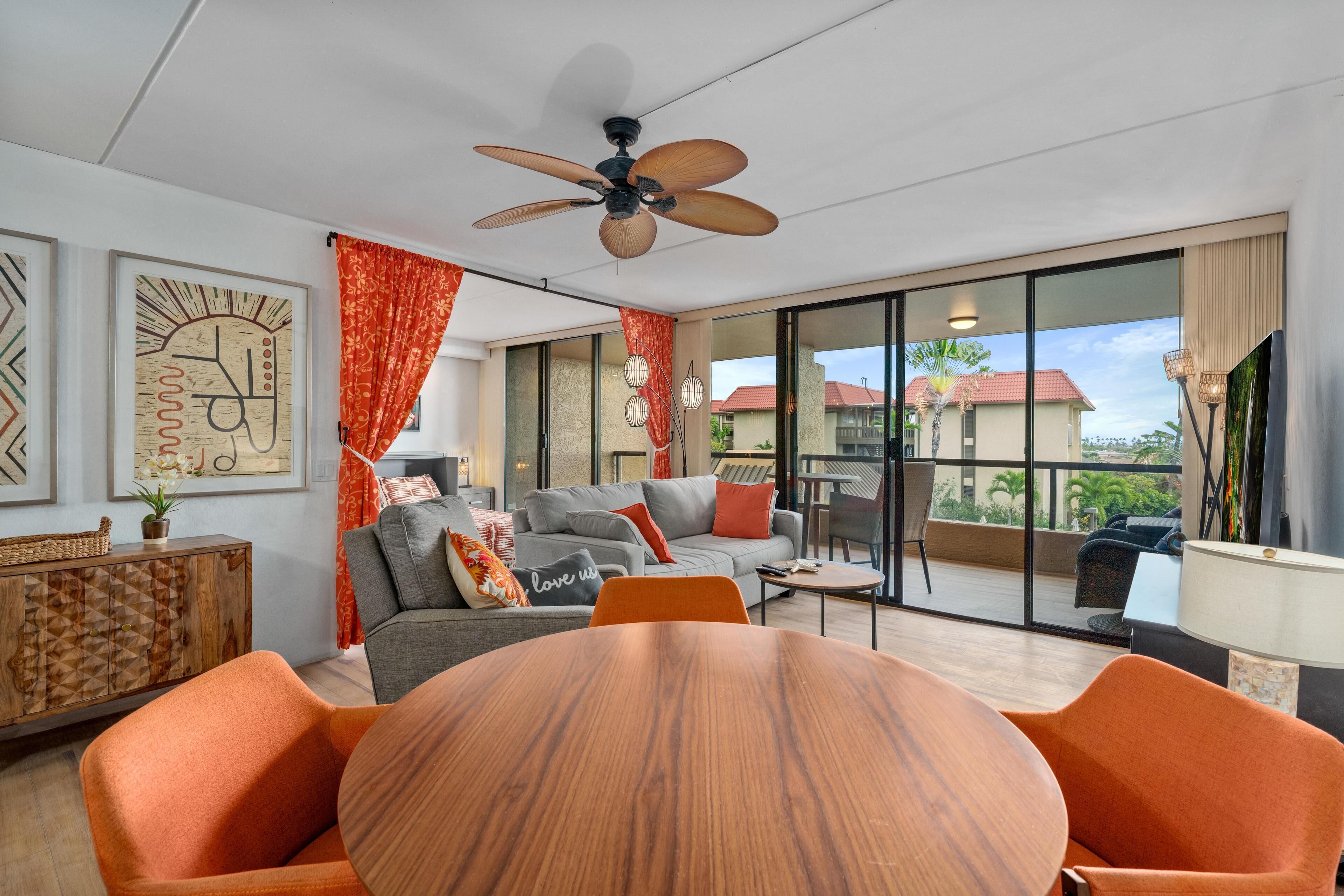 A spacious living area with a large round table, orange chairs, a grey sofa with red cushions, and a ceiling fan.