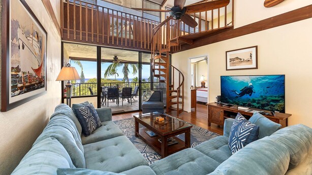 Un salón espacioso con un sofá grande, una mesa de centro y un televisor. Cuenta con un balcón con vistas a palmeras y una escalera que lleva a un nivel superior.