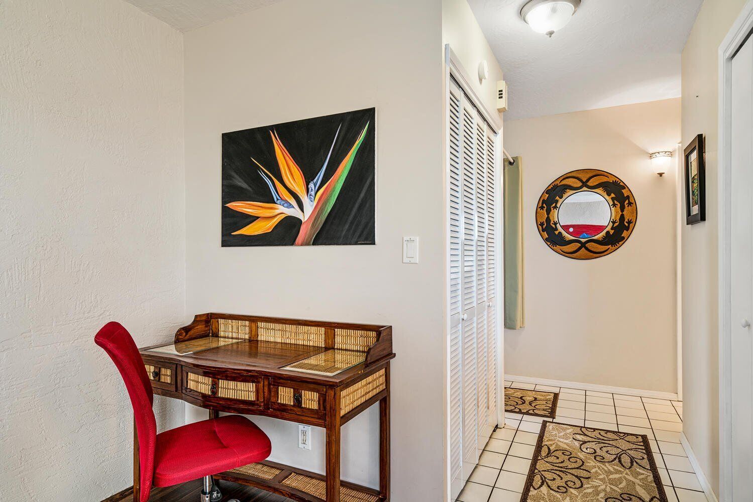 A hallway with a wooden desk, a red chair, a wall-mounted mirror, and a door with louvered shutters.