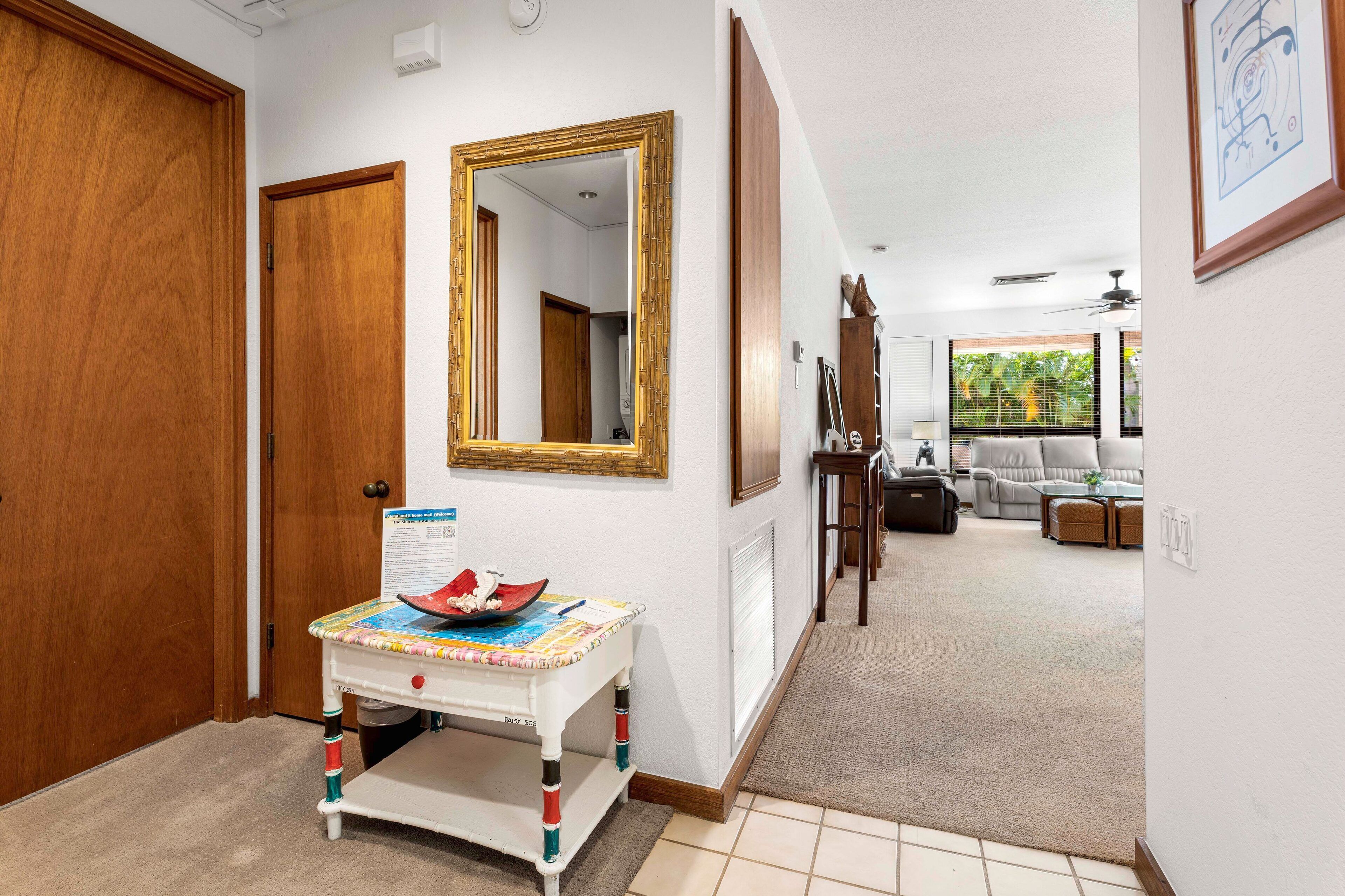 Un pasillo con una puerta de madera, un espejo, una mesita con mantel colorido y vistas a un salón con sof á y ventana.
