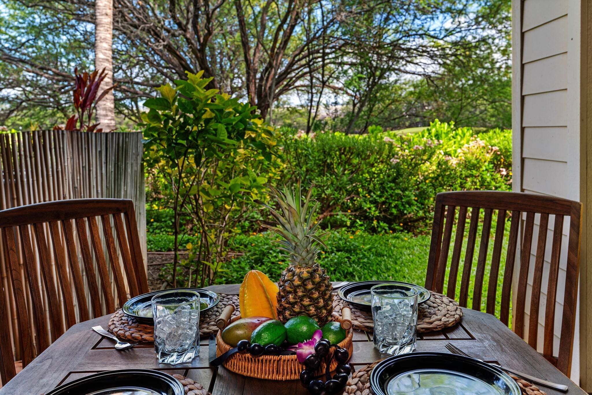 A wooden table set for two with a tropical fruit centerpiece, including a pineapple and mango, and two glasses of water.