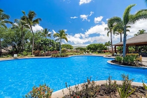 Une piscine d’un bleu éclatant, entourée de palmiers et de plantes tropicales, avec un kiosque et des chaises longues en arrière-plan.