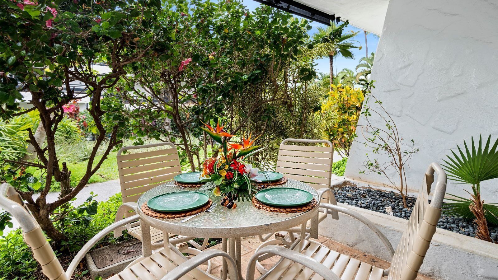Une table ronde entourée de quatre chaises, chacune agrémentée d’une assiette verte et d’une fourchette. La table est bordée d’un jardin où poussent différentes plantes, et un mur blanc se dresse en arrière-plan.