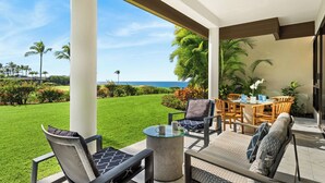 A covered patio with a view of the ocean, furnished with chairs and a table, and surrounded by greenery. - Luxury Golfside Condo w/ Ocean Views, Central AC & Beach Club - Vrkcv (Kamuela)