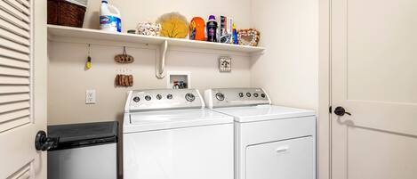 A laundry room with two washing machines, a dryer, and a shelf with various items.
