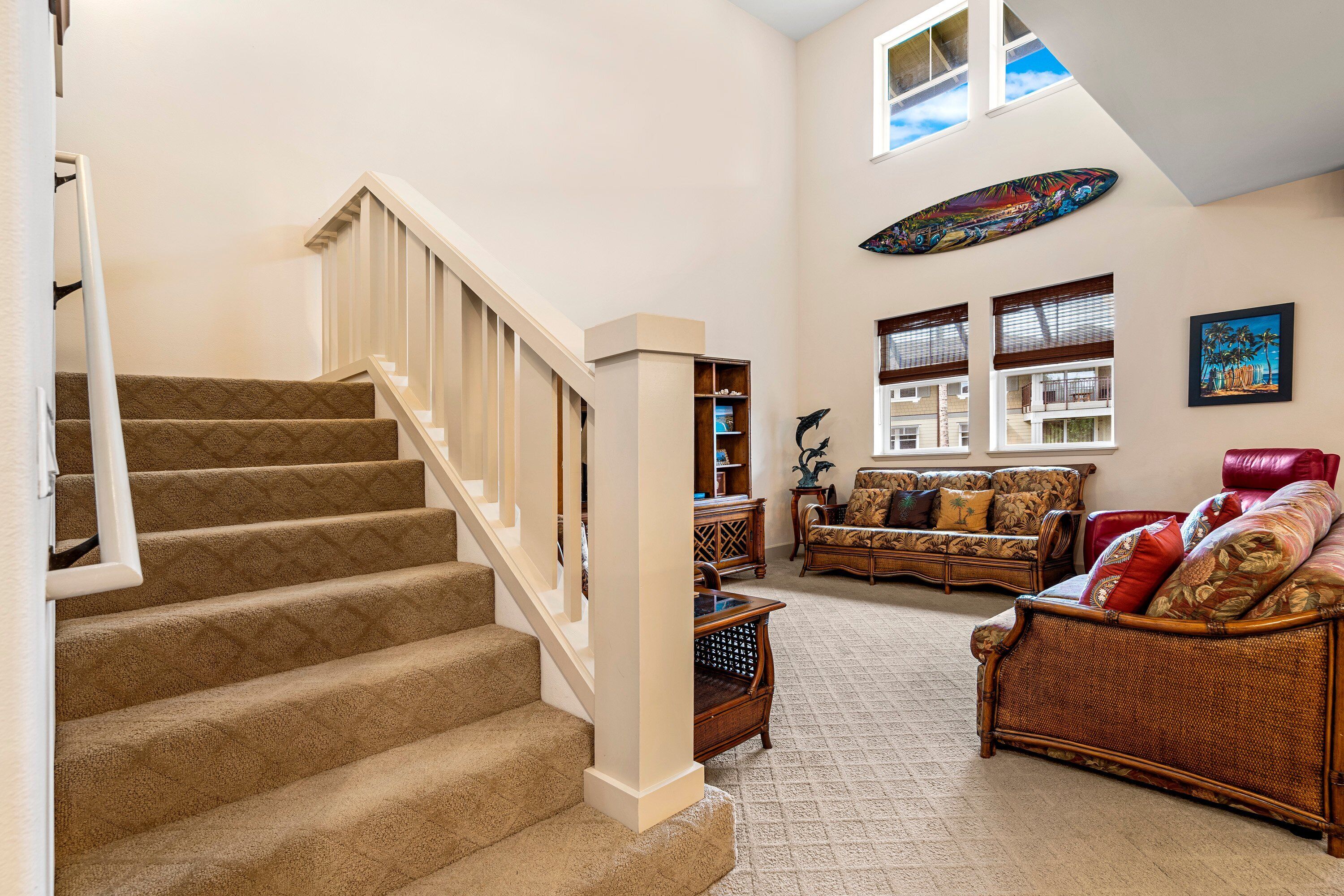 A spacious living area with a carpeted staircase, wicker furniture, and a large window with blinds.