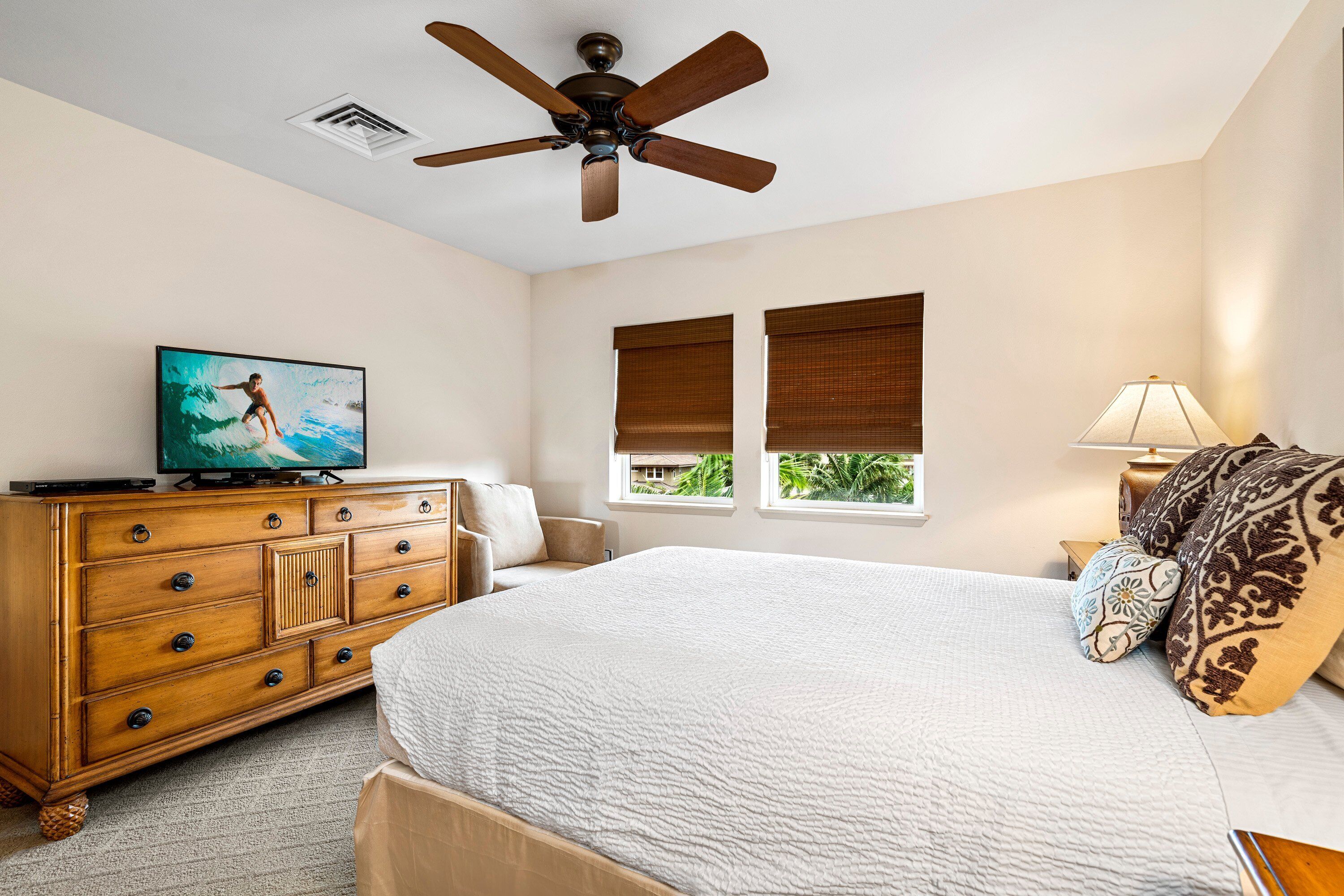 Ein Schlafzimmer mit Bett, Schminktisch, Fernseher, Deckenventilator, Fenstern und einem Nachttisch.
