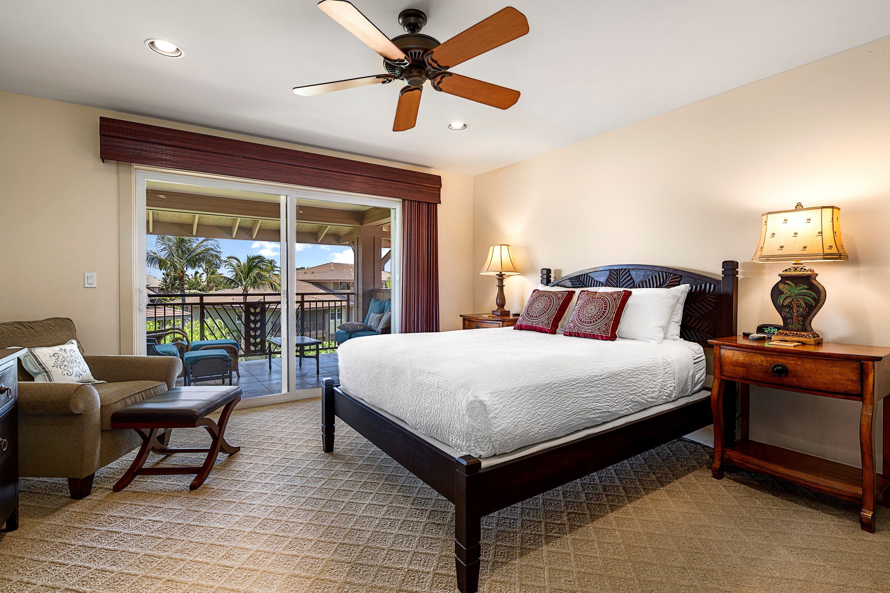 A hotel room with a large bed, a ceiling fan, a desk with a lamp, a chair, and a view of the outdoors through a sliding door.