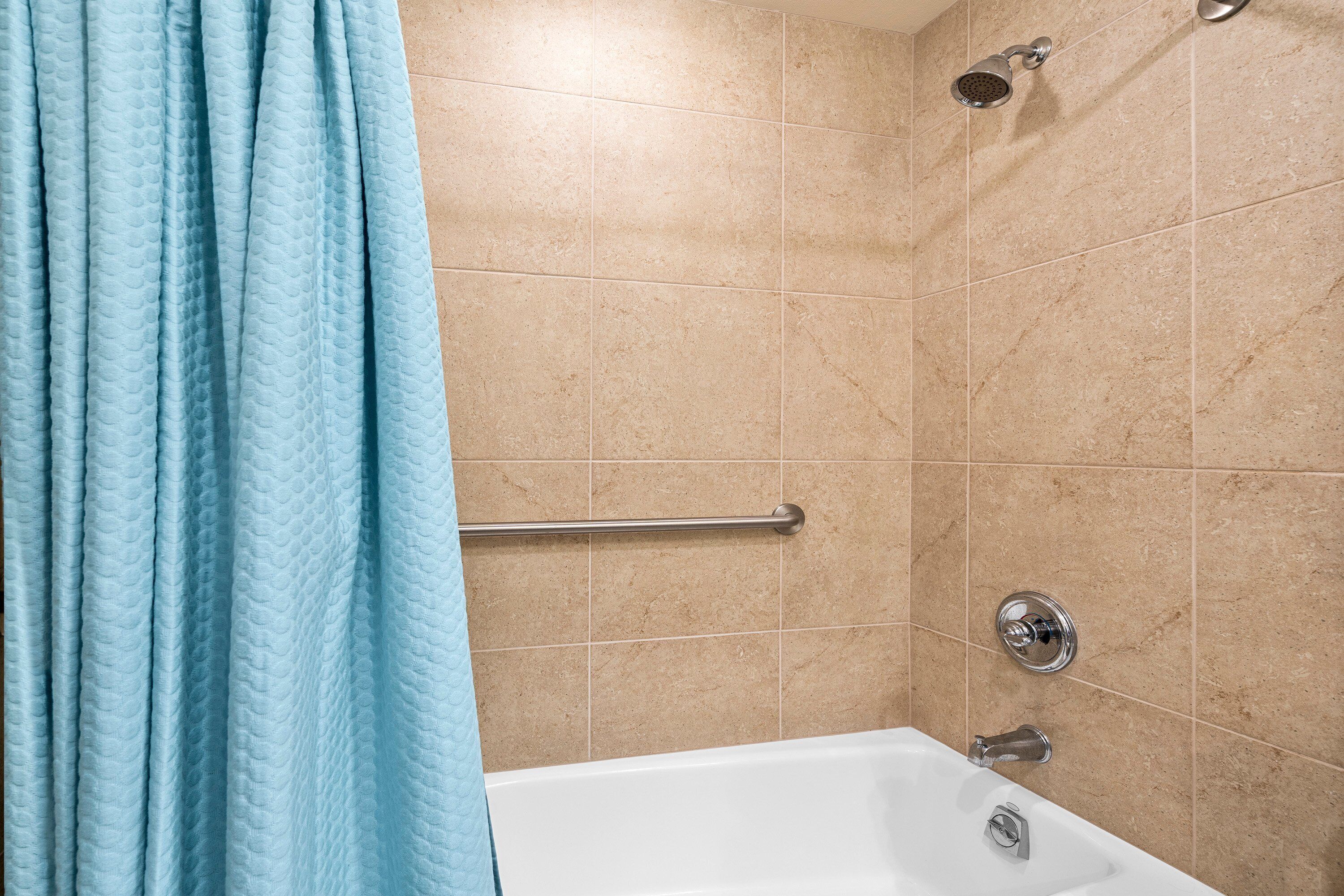 A bathroom with beige tiles, a white bathtub, a showerhead, and a grab bar.