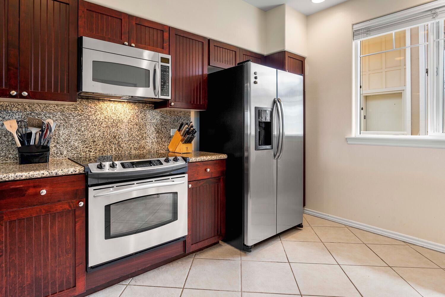 A kitchen with a stainless steel refrigerator, microwave, stove, and oven, wooden cabinets, granite countertop, and a window with blinds.