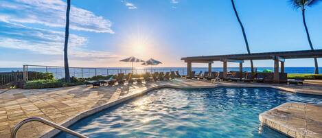 A swimming pool with sunlit water, lounge chairs, and palm trees.