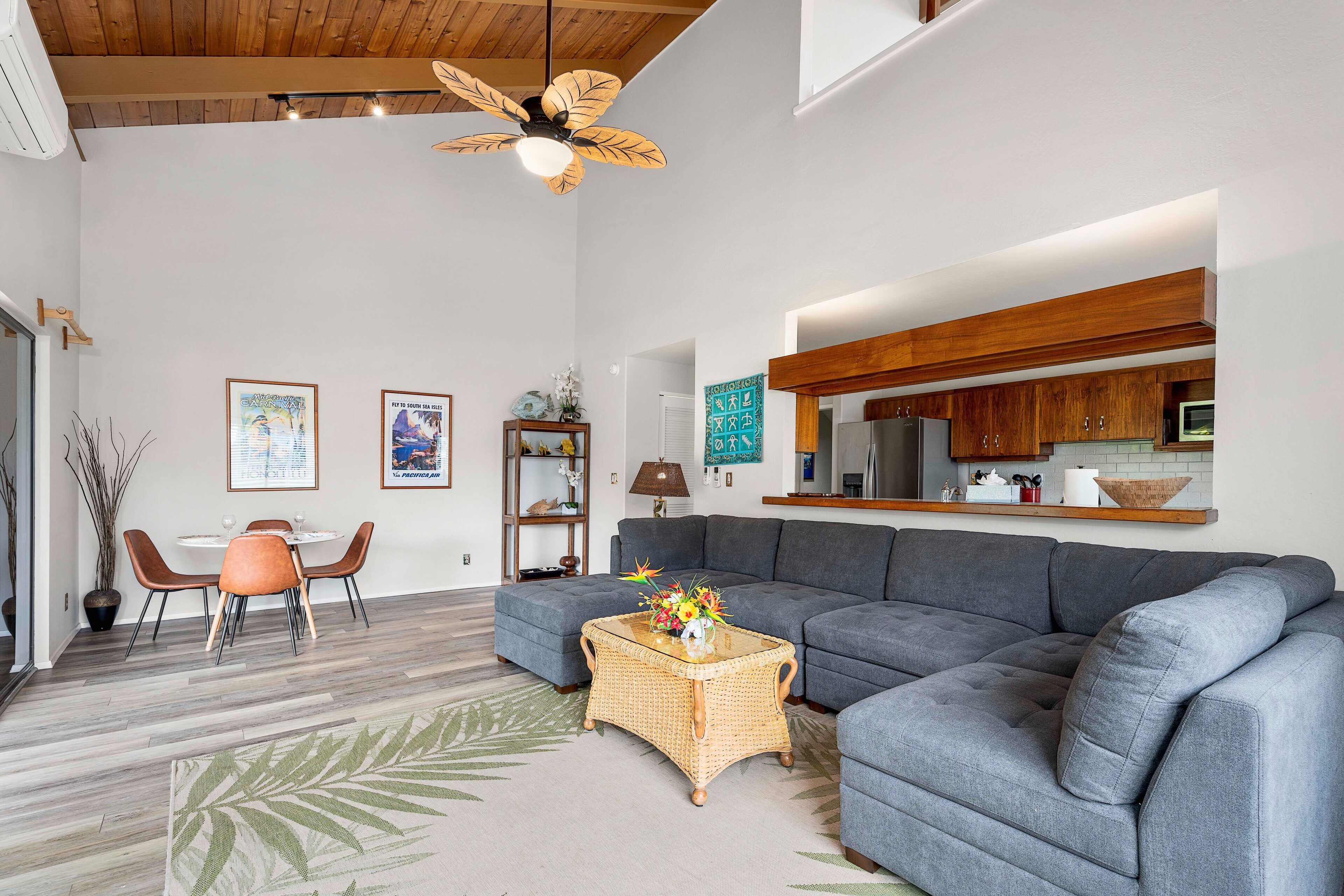 A modern living room with a grey sectional sofa, a dining table with chairs, a wicker coffee table, and a ceiling fan.