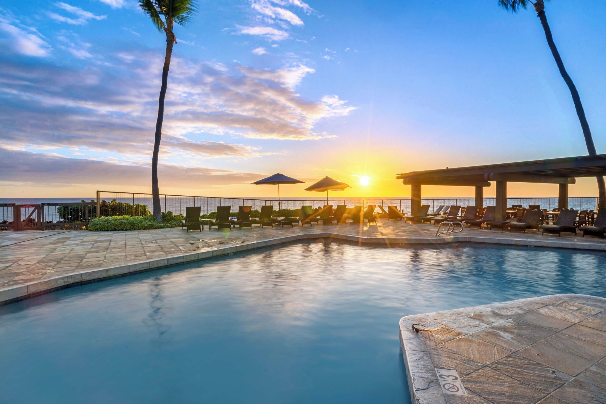 A swimming pool with lounge chairs and umbrellas, with a sunset in the background.