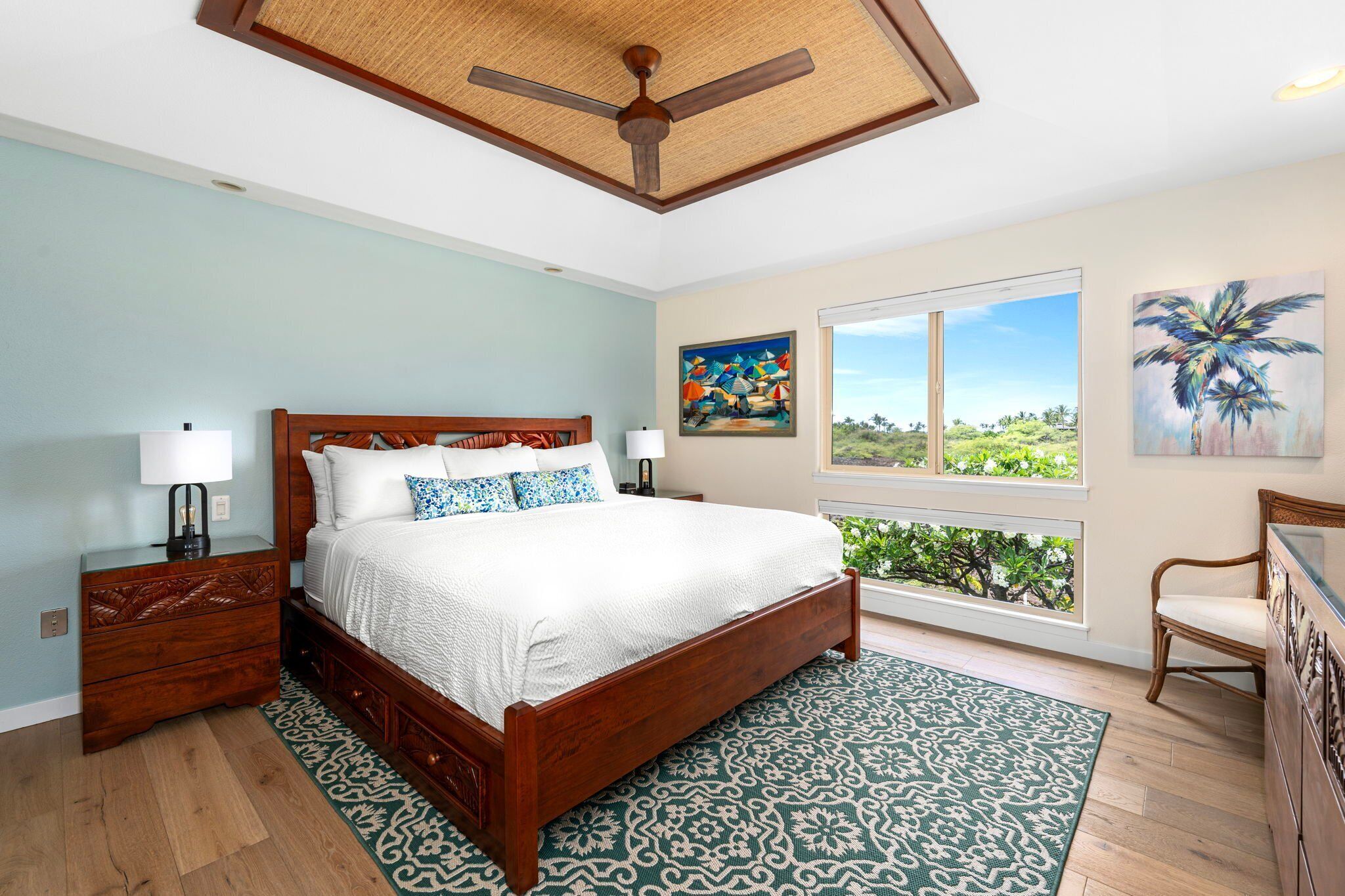 Un dormitorio con cama de madera, mesitas de noche, ventilador de techo, ventana grande con vistas y una silla.