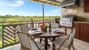 Ein Balkon mit rundem Tisch, Korbstühlen, einem Grill und Blick auf eine grüne Landschaft.