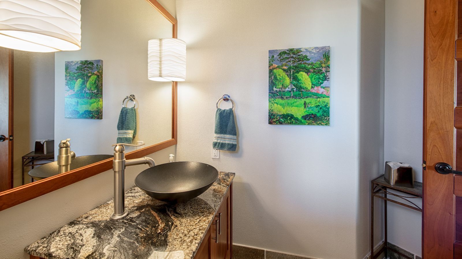 A bathroom with a large mirror, a black bowl sink, a towel rack with two towels, and two paintings on the wall.